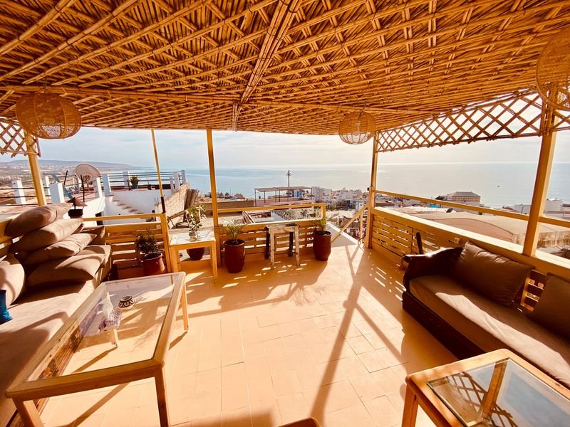 Roof terrace of the Taghazout hostel under a reed pergola: sofas, a low table and the Atlantic beyond the village