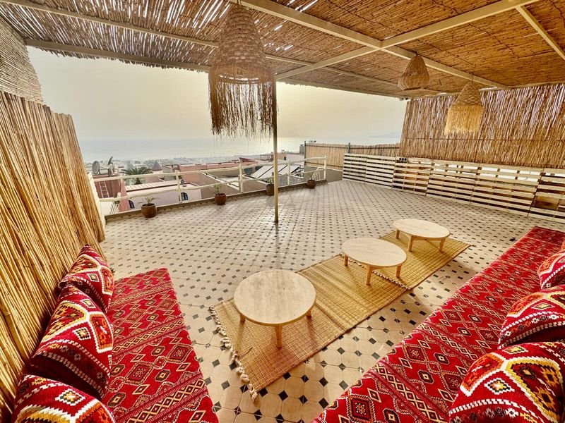 Roof terrace of the Tamraght hostel: red Berber cushions, low round tables and a reed screen, the sea in the distance